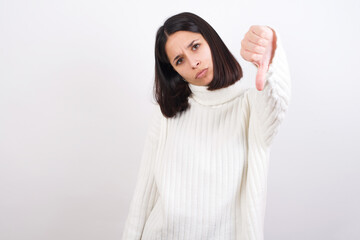 Young brunette woman wearing white knitted sweater against white background feeling angry, annoyed, disappointed or displeased, showing thumbs down with a serious look