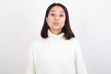 Fototapeta premium Shot of pleasant looking Young brunette woman wearing white knitted sweater against white background, pouts lips, looks at camera, Human facial expressions