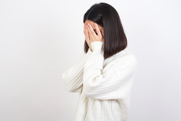 Sad Young brunette woman wearing white knitted sweater against white background crying covering her face with her hands.