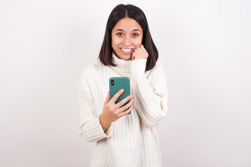 Fototapeta premium Joyous Young brunette woman wearing white sweater poses with mobile phone device, types text message on modern smartphone, watches funny video during free time, enjoys good internet connection,