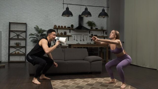 A personal trainer yells into a megaphone, making the girl do squats. Male fitness coach shouting on megaphone to motivate his female client. Motivation, workout, healthy lifestyle concept 4k
