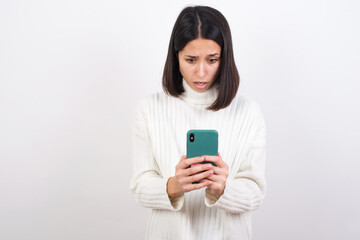 Fototapeta premium Focused Young brunette woman wearing white knitted sweater against white background use smartphone reading social media news, or important e-mail