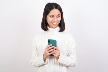 Young brunette woman wearing white knitted sweater against white background hold telephone hands read good youth news look empty space advert