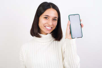 Smiling Young brunette woman wearing white knitted sweater against white background Mock up copy space. Hold mobile phone with blank empty screen