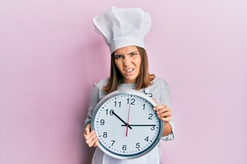 Young beautiful woman wearing professional cook uniform and hat holding clock clueless and confused expression. doubt concept.