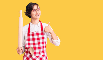 Beautiful young woman with short hair wearing professional baker apron holding kneading roll smiling happy and positive, thumb up doing excellent and approval sign