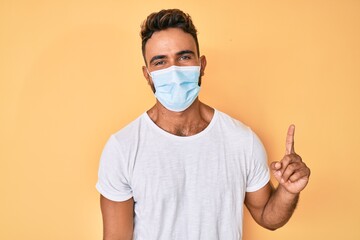 Young hispanic man wearing medical mask smiling with an idea or question pointing finger up with happy face, number one