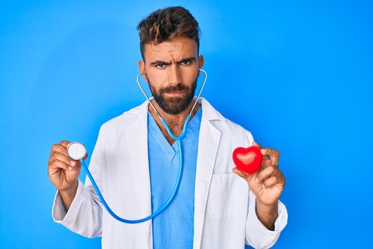 Young hispanic man wearing doctor uniform holding stethoscope and heart skeptic and nervous, frowning upset because of problem. negative person.