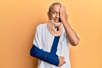 Handsome mature senior man wearing cervical collar and arm on sling covering one eye with hand,...