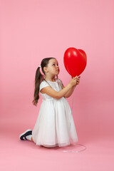 a pensive, dreamy girl with ponytails and a white dress is kneeling and looking at a red balloon in the shape of a heart , isolated on a pink background.