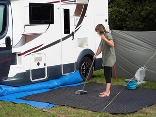 A lady motorhome owner sweeps her outdoor mat with a broom outside her recreational vehicle. A...