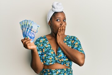 Young african woman wearing hair turban holding south african 100 rand banknotes covering mouth...