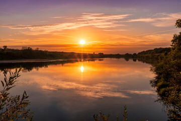 Scenic view at beautiful summer river sunset with reflection on water with green bushes, grass, golden sun rays, calm water ,deep blue cloudy sky and glow on a background, spring evening landscape