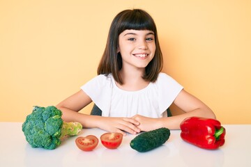 Young little girl with bang sitting on the table with veggies with a happy and cool smile on face. lucky person.
