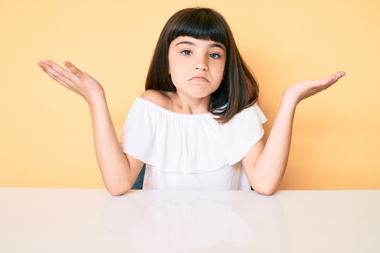 Young little girl with bang wearing casual clothes sitting on the table clueless and confused with open arms, no idea and doubtful face.