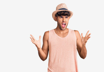 Young hispanic man wearing summer hat crazy and mad shouting and yelling with aggressive expression and arms raised. frustration concept.