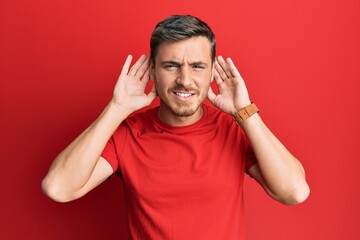 Handsome caucasian man wearing casual red tshirt trying to hear both hands on ear gesture, curious for gossip. hearing problem, deaf