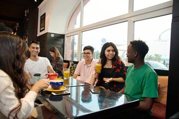 Multiracial happy young people drink coffee at cafe, black and white cheerful mates laughing enjoying drinks having fun sitting together at restaurant table, diverse friends share lunch at meeting