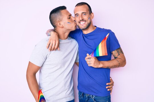 Young couple of gay men together in love holding homosexual pride colorful flag kissing with affection