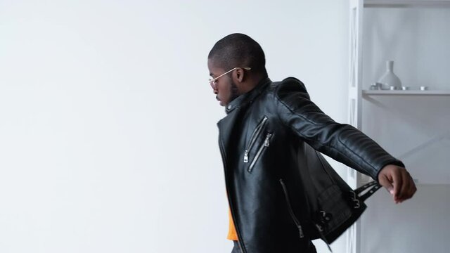 African man lifestyle. Black lives matter. Stylish masculine confident dark skin guy wearing leather bicker jacket orange cap sunglasses sitting down looking at camera on light copy space background.