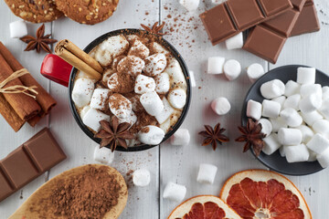 Red mug of hot chocolate with marshmallow, anise and cinnamon sprinkled with cocoa powder on a wooden table with slices of dried grapefruit and chocolate pieces. Winter hot drink. Top view.