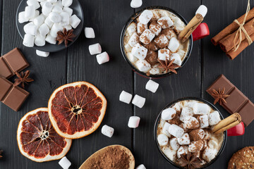 Fototapeta premium Two red cups of hot chocolate with marshmallow, anise and cinnamon sprinkled with cocoa powder on a wooden table with slices of dried grapefruit and chocolate pieces. Winter hot drink top view.