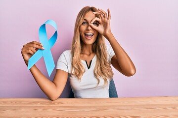 Beautiful blonde young woman holding blue ribbon smiling happy doing ok sign with hand on eye...