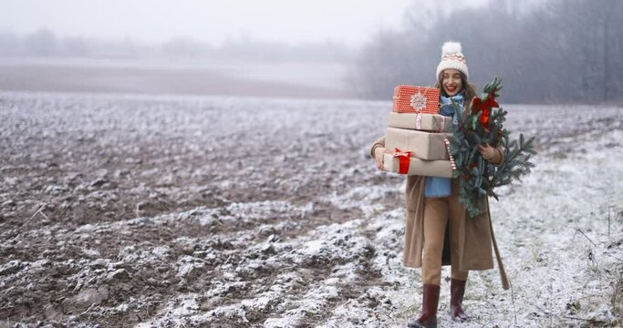 Happy woman walking with gift boxes and Christmas branch on the snowy field outdoors. Concept of a festive mood, lifestyle and celebrating in countryside