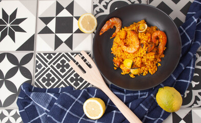 Mesa tradicional con un plato negro de arroz en paella, limones alrededor del plato,una servilleta azul y un tenedor.