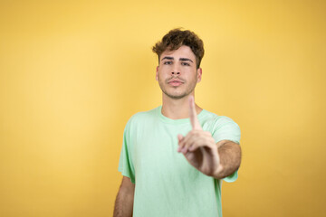 Handsome man wearing a green casual t-shirt over yellow background showing and pointing up with fingers number one while is serious