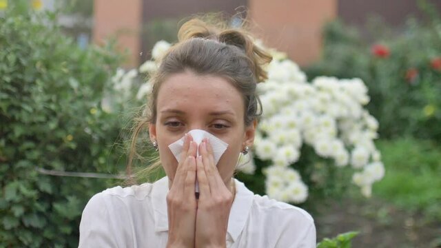 Girl sneezes into a napkin, because she is allergic to flowering during spring time on white chrysanthemums on background, hypersensitivity concept