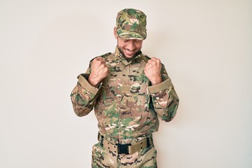 Young caucasian man wearing camouflage army uniform excited for success with arms raised and eyes...