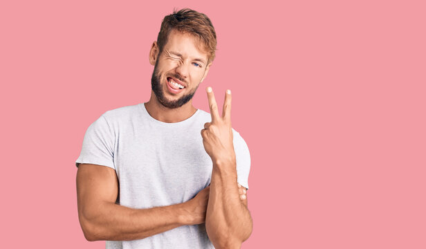 Young caucasian man wearing casual white tshirt smiling with happy face winking at the camera doing victory sign. number two.