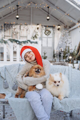 Joyful child, a cute boy dressed in a red santa claus hat holds two Spitz puppies in his hands on the background of a white house in winter. Christmas gift for a child - 402911389