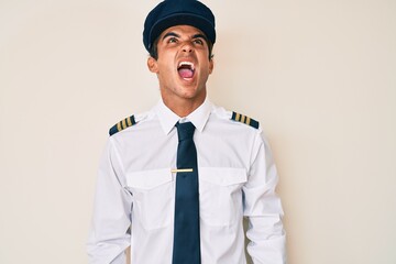 Young hispanic man wearing airplane pilot uniform angry and mad screaming frustrated and furious, shouting with anger. rage and aggressive concept.