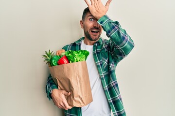 Handsome man with beard holding paper bag with groceries surprised with hand on head for mistake,...