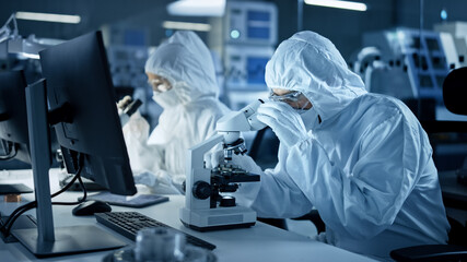 Research Factory Cleanroom: Engineers / Scientists wearing Coveralls and Gloves Use Microscopes to...