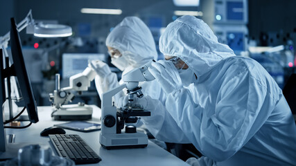 Research Factory Cleanroom: Engineers / Scientists wearing Coveralls and Gloves Use Microscopes to...