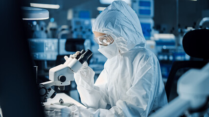 Research Factory Cleanroom: Engineer / Scientist wearing Coverall and Gloves Uses Microscope to...