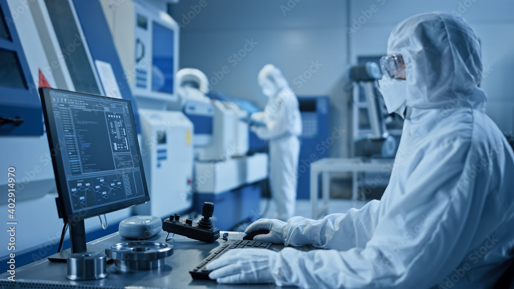 Poster Factory Cleanroom: Engineer Wearing Coverall and Mask Does System Control on a Computer, Uses Controller, Screen Shows Infographics. In Background Modern CNC Machinery, Robot Arm, Electronic Equipment