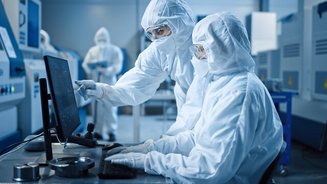 Factory Cleanroom: Engineer and Scientist Wearing Coveralls and Masks Have Discussion, Use Computer Showing Infrastructure System Control. In Background CNC Machinery, Electronics Equipment