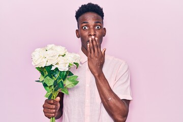 Young african american man holding flowers covering mouth with hand, shocked and afraid for...