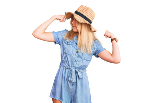 Young beautiful blonde woman wearing summer hat and dress showing arms muscles smiling proud. fitness concept.