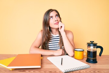 Beautiful young caucasian woman sitting on the table stuying for university thinking concentrated about doubt with finger on chin and looking up wondering