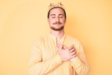 Young handsome man wearing king crown smiling with hands on chest, eyes closed with grateful...