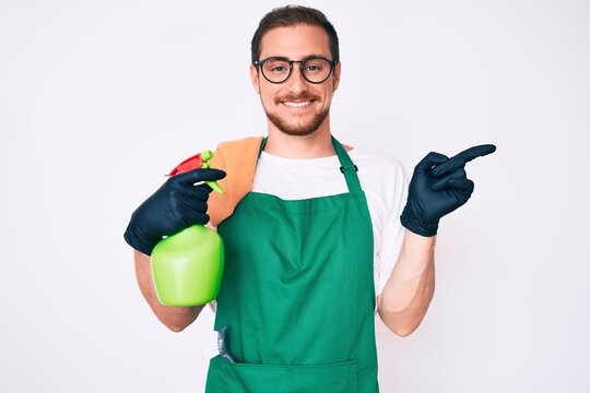 Young handsome man wearing apron holding sprayer smiling happy pointing with hand and finger to the side