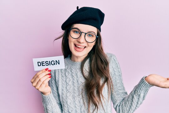 Young beautiful caucasian girl holding design word on paper celebrating achievement with happy smile and winner expression with raised hand