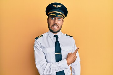 Handsome hispanic man wearing airplane pilot uniform pointing aside worried and nervous with...