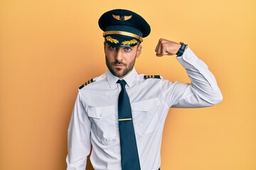 Handsome hispanic man wearing airplane pilot uniform strong person showing arm muscle, confident and proud of power