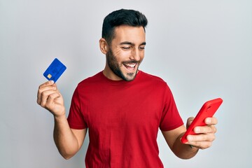 Young hispanic man holding smartphone and credit card smiling and laughing hard out loud because funny crazy joke.